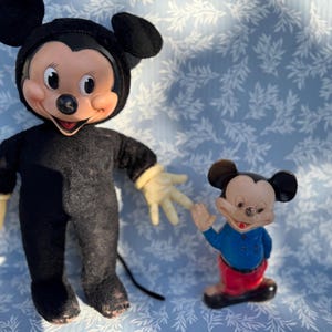 May include: Two vintage Mickey Mouse figures. One is a plush doll in a black outfit with yellow gloves. The other is a small figurine wearing a blue shirt, red pants, and black shoes. Both have the iconic Mickey Mouse face.