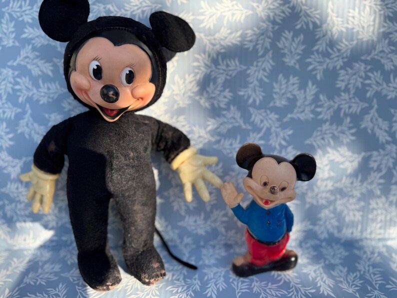 May include: Two vintage Mickey Mouse figures. One is a plush doll in a black outfit with yellow gloves. The other is a small figurine wearing a blue shirt, red pants, and black shoes. Both have the iconic Mickey Mouse face.