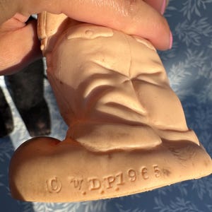 May include: A vintage, peach-colored figurine of a character in a uniform. The figurine is held by a hand, with the bottom of the figurine visible, displaying the copyright mark and the text "&copy; W.D.P. 1965".