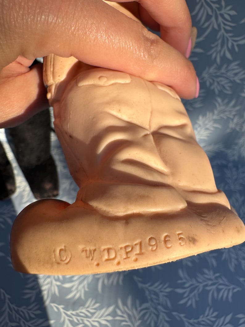 May include: A vintage, peach-colored figurine of a character in a uniform. The figurine is held by a hand, with the bottom of the figurine visible, displaying the copyright mark and the text "&copy; W.D.P. 1965".