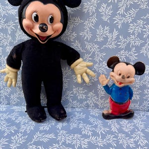 May include: Two vintage Mickey Mouse figurines. One is a plush doll in a black outfit with a smiling face and cream-colored hands. The other is a small plastic figurine of Mickey Mouse in a blue shirt and red pants, waving.