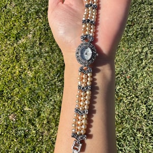May include: A vintage-style wristwatch with a pearl bracelet. The watch face is round with a silver bezel and black markers. The bracelet is made of multiple strands of cream-colored pearls, accented with silver-toned decorative elements.