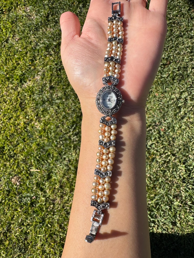May include: A vintage-style wristwatch with a pearl bracelet. The watch face is round with a silver bezel and black markers. The bracelet is made of multiple strands of cream-colored pearls, accented with silver-toned decorative elements.