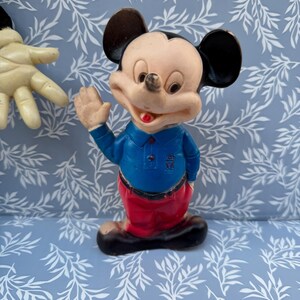 May include: A vintage Mickey Mouse figurine. The figure is wearing a blue shirt, red pants, and black shoes. Mickey has black ears, a tan face, and is waving with his right hand. The figurine is on a blue background.
