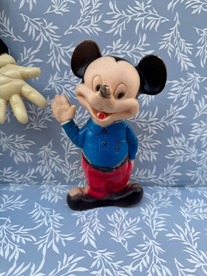 May include: A vintage Mickey Mouse figurine. The figure is wearing a blue shirt, red pants, and black shoes. Mickey has black ears, a tan face, and is waving with his right hand. The figurine is on a blue background.