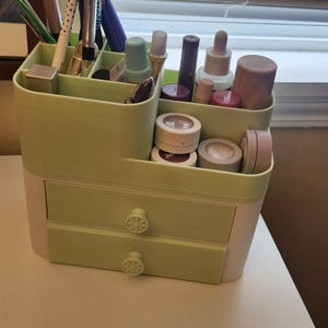 May include: A light green and white makeup organizer with two drawers and multiple compartments. The organizer is filled with various cosmetics, including lipsticks, eye shadows, and skincare products. The drawers have decorative floral knobs.