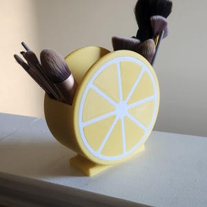 May include: A yellow, lemon-shaped makeup brush holder with white accents. The holder is filled with various makeup brushes. The design resembles a lemon slice, adding a fun and decorative touch to any vanity or bathroom.