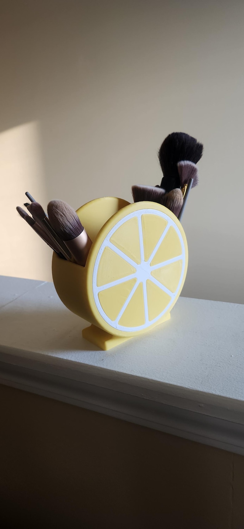 May include: A yellow, lemon-shaped makeup brush holder with white accents. The holder is filled with various makeup brushes. The design resembles a lemon slice, adding a fun and decorative touch to any vanity or bathroom.