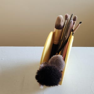 May include: A collection of makeup brushes in a yellow holder. The brushes vary in size and shape, with black, gold, and pink handles. The bristles are in shades of brown and black. The holder is on a white surface.
