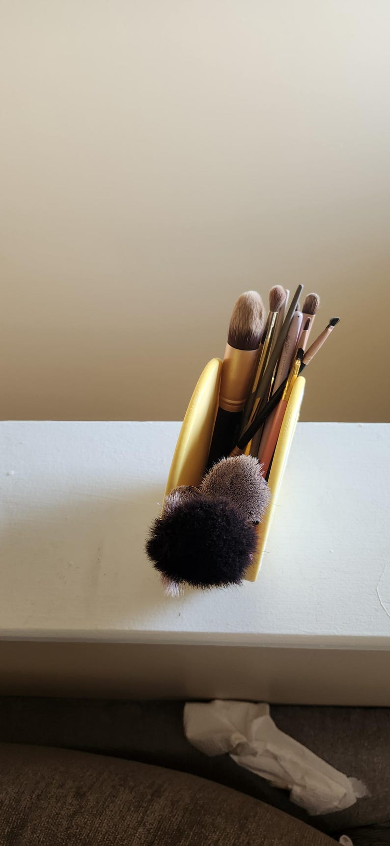 May include: A collection of makeup brushes in a yellow holder. The brushes vary in size and shape, with black, gold, and pink handles. The bristles are in shades of brown and black. The holder is on a white surface.