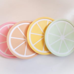 Puede incluir: Cuatro posavasos coloridos con forma de rodajas de cítricos. Los posavasos están dispuestos en fila, con colores que incluyen rosa, melocotón, amarillo y verde. Cada posavasos tiene un diseño interior blanco que imita los gajos de una fruta.