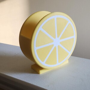 May include: A yellow lemon-shaped napkin holder with white accents. The holder is made of a solid material and features a detailed lemon slice design. The product is designed to hold napkins and is a decorative kitchen accessory.