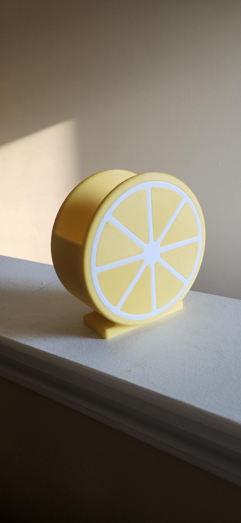 May include: A yellow lemon-shaped napkin holder with white accents. The holder is made of a solid material and features a detailed lemon slice design. The product is designed to hold napkins and is a decorative kitchen accessory.
