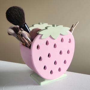 May include: A pink and green strawberry-shaped makeup brush holder with a green stem and leaves. The holder has holes for holding brushes and is filled with makeup brushes.