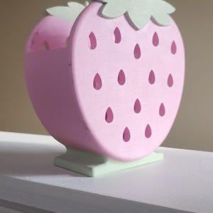 Peut inclure: Un organisateur en bois en forme de fraise rose et verte avec des d&eacute;tails d&eacute;coup&eacute;s. L'organisateur a une base plate et est parfait pour ranger de petits objets.