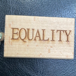 May include: A wooden keychain with the word "EQUALITY" engraved in a brown font. The keychain is rectangular and attached to a silver metal ring.