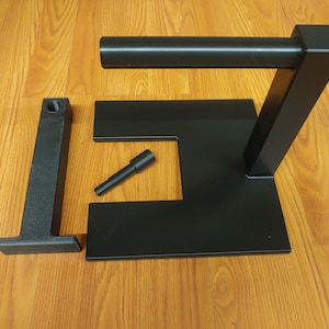 May include: Black metal stand with a square base and a vertical post with a horizontal arm. The stand is disassembled and includes a small black metal pin.