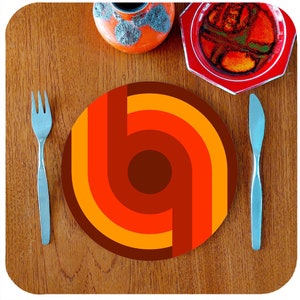 May include: A round placemat with a geometric design in shades of orange, red, and brown. The placemat has a concentric circle pattern with a large brown circle in the center.