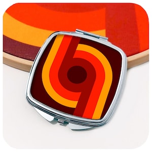May include: A compact mirror with a retro design featuring a red, orange, and yellow geometric pattern. The mirror has a silver frame.