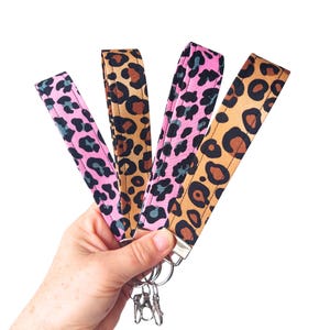 May include: Four leopard print wristlet keychains in pink, tan, and brown. Each keychain has a different colour scheme, with silver-tone hardware and key rings. The keychains are held in a hand against a white background.