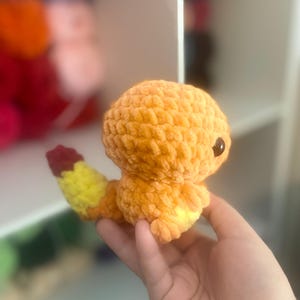 May include: A handmade orange crochet creature with a yellow and red tail. The plush toy has a round head, small arms and legs, and a dark eye. The toy is held in a hand.