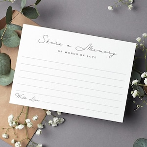 May include: A white card with the words "Share a Memory or Words of Love" in elegant script. The card has lined spaces for writing and the words "With Love" at the bottom. The card is surrounded by greenery and small white flowers.