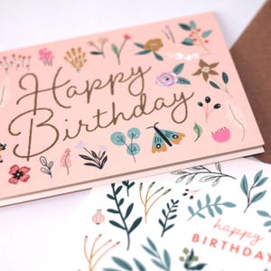 May include: A pink birthday card with the text "Happy Birthday" in gold lettering. The card is decorated with a floral pattern in shades of pink, green, and brown.