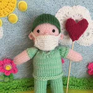 Może przedstawiać: Pluszowa lalka ubrana w zielony fartuch i białą maskę na twarz, trzymająca czerwone serce. Lalka ma zielony kapelusz i różową twarz.