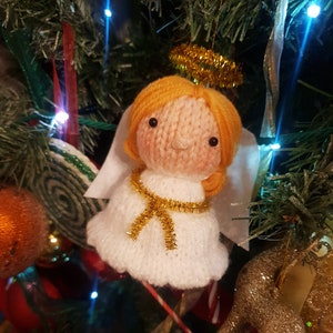 May include: A knitted angel ornament with a white dress, gold halo, and gold ribbon. The angel has light orange hair and a sweet expression. The ornament is hanging on a Christmas tree.