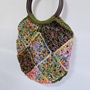 May include: A handmade crochet bag with a wooden handle. The bag features a patchwork design in shades of green, pink, yellow, and blue. The handle is a dark brown, and the bag is suspended against a white background.