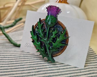 Puede incluir: Broche artesanal con un diseño detallado de cardo. El broche presenta una vibrante flor morada y hojas verdes, sobre una base de madera. El broche está sobre una tarjeta blanca, con una tela a rayas en el fondo.