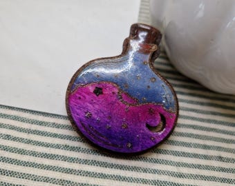 May include: A wooden pendant in the shape of a potion bottle. The bottle features a gradient of purple and blue with a starry night design, including a crescent moon and a star. The bottle has a brown wooden frame and a brown cork.