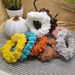 May include: A collection of colorful, fluffy scrunchies in various shades including yellow, blue, orange, brown, pink, white, and gray. The scrunchies are made of a soft, textured material. A white pumpkin and a small cactus in a white pot are in the background.