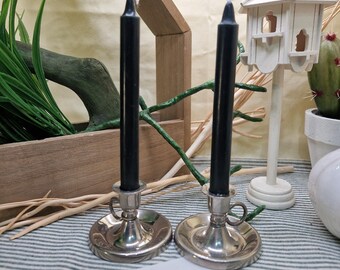 May include: Two black taper candles in silver-colored candle holders. The candle holders have a round base and a small handle. The candles are unlit and stand upright. The background includes a wooden birdhouse and decorative greenery.