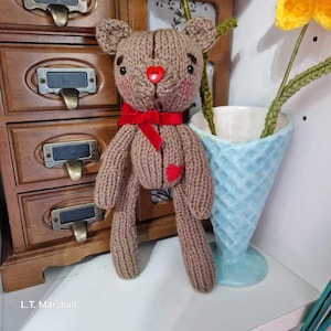 May include: A brown knitted teddy bear with a red heart on its chest and a red bow around its neck. The bear is standing on a white surface.