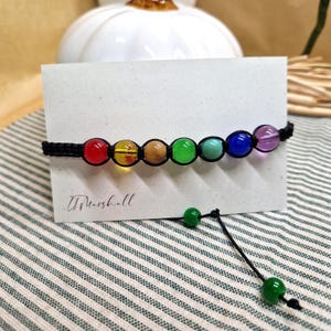 Puede incluir: Una pulsera hecha a mano con una serie de cuentas redondas y coloridas en un espectro de arcoíris, ensartadas en un cordón negro. Las cuentas incluyen rojo, amarillo, marrón, verde, turquesa, azul y morado. La pulsera se muestra en una tarjeta blanca.