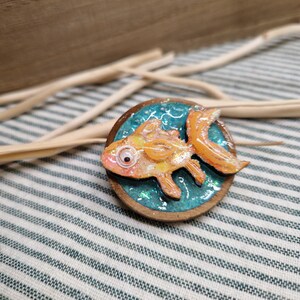 以下が含まれることがあります： 金魚のデザインが施された手作りの木製ピンバッジ。オレンジと黄色の金魚は、キラキラ光るターコイズブルーの背景にセットされ、円形の木製フレームの中に収められています。ユニークなアクセサリーです。