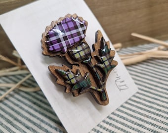 Puede incluir: Broche de madera con forma de flor de cardo. La cabeza de la flor presenta un estampado de cuadros morados y blancos, mientras que las hojas y el tallo tienen un diseño de cuadros verdes y amarillos. El broche está sobre una tarjeta blanca.