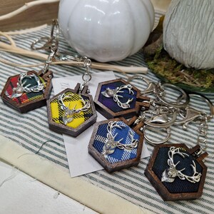 May include: Keychains with a hexagonal wooden frame and a silver deer head emblem. The keychains feature colorful plaid fabric inserts in various colors, including yellow, green, blue, and purple. Each keychain has a silver-colored clasp.