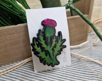 Puede incluir: Broche de cardo con una flor rosa brillante, tallo verde y hojas verdes con contornos negros. El broche está montado en una tarjeta blanca. El fondo incluye una caja de madera, vegetación y tela a rayas.