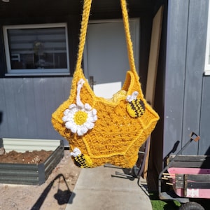 Könnte beinhalten: Handgefertigte, gelbe Häkeltasche in Sternform. Sie hat eine weiße und gelbe Gänseblümchenblume und drei aufgenähte Bienen. Der Träger ist ebenfalls gehäkelt. Die Tasche hängt im Freien im natürlichen Licht.