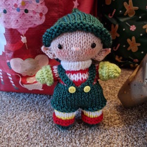 May include: A handmade knitted elf doll with a green hat and overalls. The doll has a white and red striped shirt, green mittens, and yellow buttons. The elf's shoes are red, yellow, and white striped. The doll is approximately 6 inches tall.