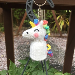 Può includere: Un portachiavi a forma di unicorno di lana bianca con una criniera e un corno arcobaleno. Il portachiavi è appeso a un gancio di metallo.