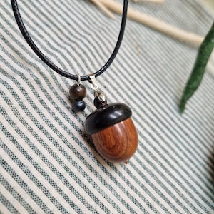 Op de afbeelding: Een houten eikelhangerketting met een zwart koord. De eikelhanger heeft een donkerbruine dop en een lichtbruin lichaam. Twee kleine houten kralen hangen aan het koord bij de hanger. De ketting rust op een gestreepte stof.