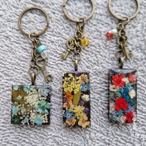 May include: Three keychains with different charms and resin squares containing pressed flowers. The charms include keys, hearts, and beads. The resin squares are in shades of blue, red, and yellow.