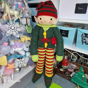May include: A knitted elf doll with a green sweater, red and yellow striped pants, and a red hat with white stripes. The elf is standing next to a white Christmas tree.