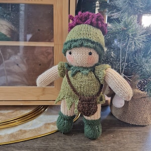 May include: A handmade knitted doll with a whimsical design. The doll is dressed in a green outfit with a matching hat and boots. It has a small brown bag and a pink flower on top of its head. The doll is standing in front of a Christmas tree.