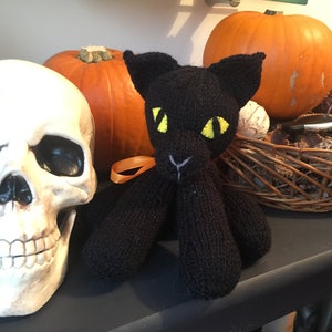 May include: A black knitted cat toy with yellow eyes sits on a table with a white plastic skull and two orange pumpkins.