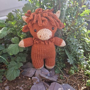 May include: A brown and cream knitted stuffed toy cow with a fluffy brown mane. The toy is standing on a bed of small grey rocks.
