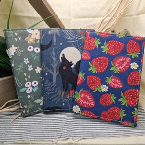 May include: Three fabric-covered notebooks. One has a floral pattern in green, white, and beige. Another features a wolf howling at the moon in a forest scene. The third notebook has a pattern of red strawberries on a blue background.